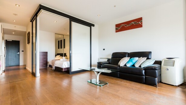 A modern living room with a glass coffee table, a black leather sofa with cushions, a white air conditioner, and a sliding glass door leading to another room. - Cleo, 1BDR St Kilda Apartment (St Kilda)