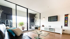 A modern living room with a glass coffee table, a black leather sofa, a TV mounted on the wall, and a sliding glass door leading to a balcony. - Cleo, 1BDR St Kilda Apartment (St Kilda)