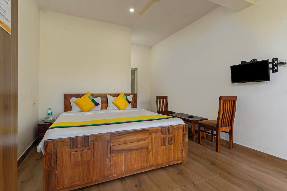Uma cama de madeira com colcha colorida, uma televisão presa na parede, uma cadeira de madeira e uma mesinha.
