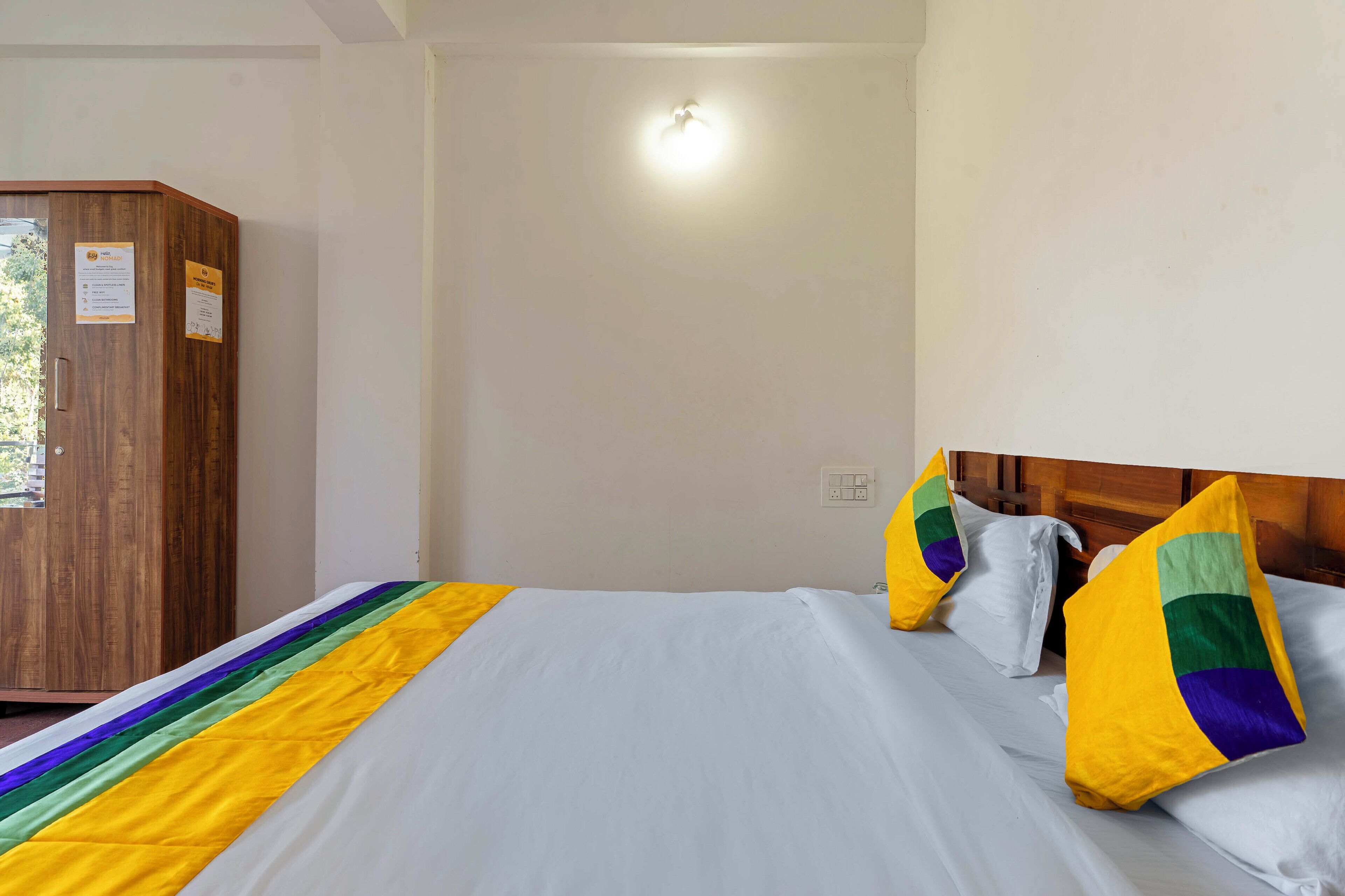 Una habitación de hotel con un armario de madera, una cama con ropa de cama blanca y almohadas de colores, e iluminación en el techo.