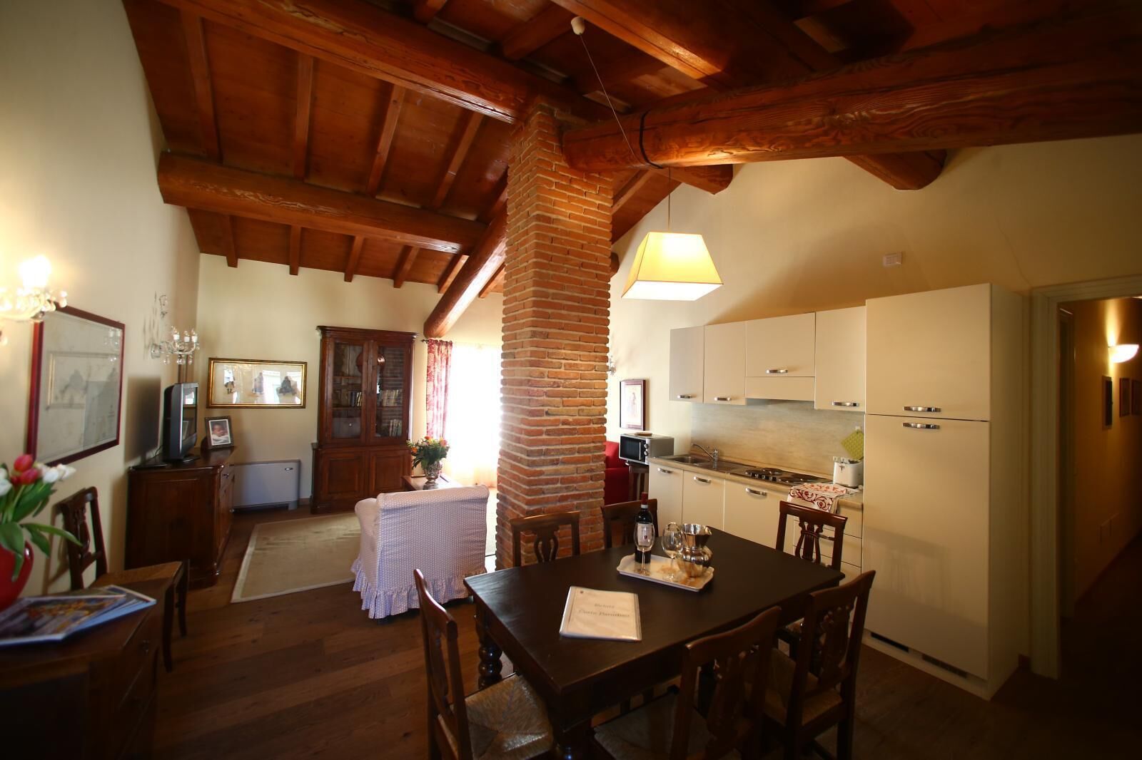 Un coin repas chaleureux avec une table et des chaises en bois, un pilier en briques et une cuisine dotée de meubles blancs et d’un réfrigérateur.