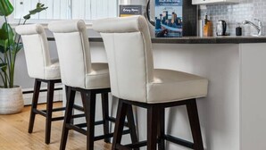 A modern kitchen with white cabinetry, a built-in island with bar stools, and a large window with blinds. - Bright and Open Coziness. 10 minutes to Stampede (Calgary)