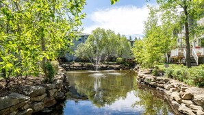 A tranquil pond with a fountain, surrounded by stone walls and lush greenery, with residential buildings in the background. - Bright and Open Coziness. 10 minutes to Stampede (Calgary)