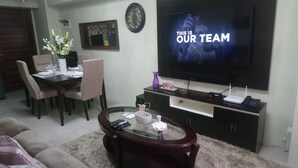 Una sala de estar con un televisor mostrando 'THIS IS OUR TEAM', un comedor con sillas y una mesa, y una mesa de centro con controles remotos.