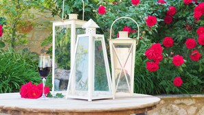 A garden table with two white lanterns, a glass of red wine, and a red flower. - Niko's Art villa (Chania)