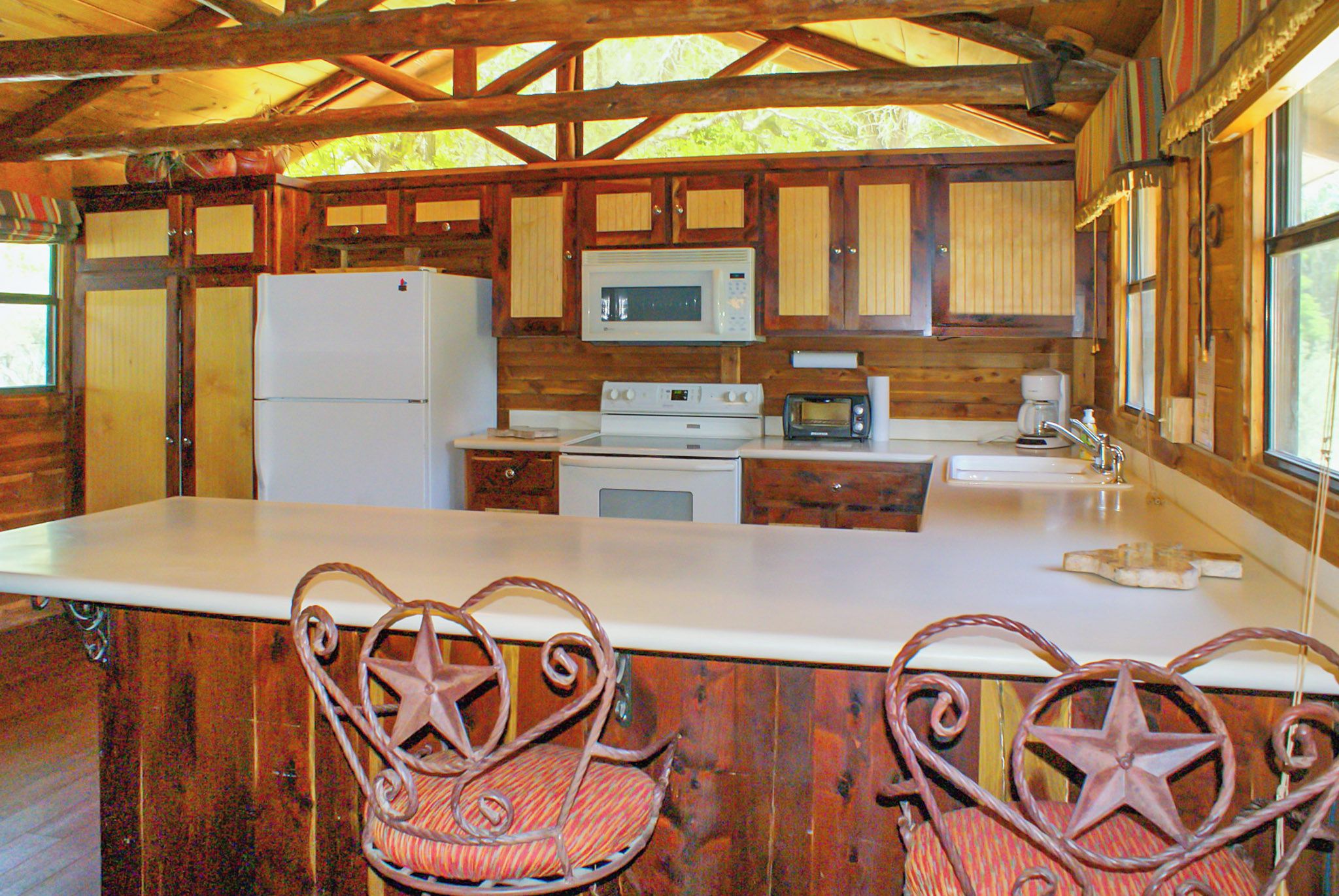 A rustic cabin kitchen with wooden cabinets, a white refrigerator, a microwave, a stove, and two star-shaped chairs.