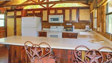 A rustic cabin kitchen with wooden cabinets, a white refrigerator, a microwave, a stove, and two star-shaped chairs.