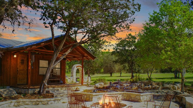 Una cabaña rústica con chimenea de piedra y sillas metálicas, rodeada de un patio de piedra y vista al atardecer.