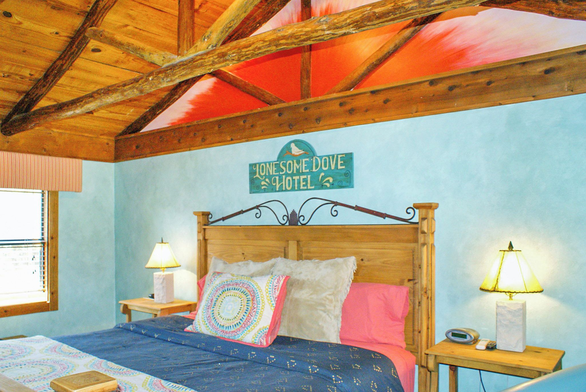 Ett sovrum med ett träpanelat tak, en säng med mönstrat sänglinne, två sängbord med lampor och en skylt ovanför sängen med texten ”Lonesome Dove Hotel”.