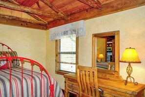 Una habitación con cama, una silla de madera, una mesa con lámpara, una ventana con persianas y un espejo en la pared.