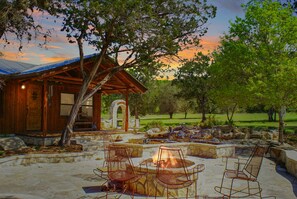 A rustic cabin with a stone fireplace and metal chairs, surrounded by a stone patio and a view of the sunset. - Charming Creekside Cabin w/ Hot Tub, Fire Pit & Natural Rock Waterfalls (Wimberley)