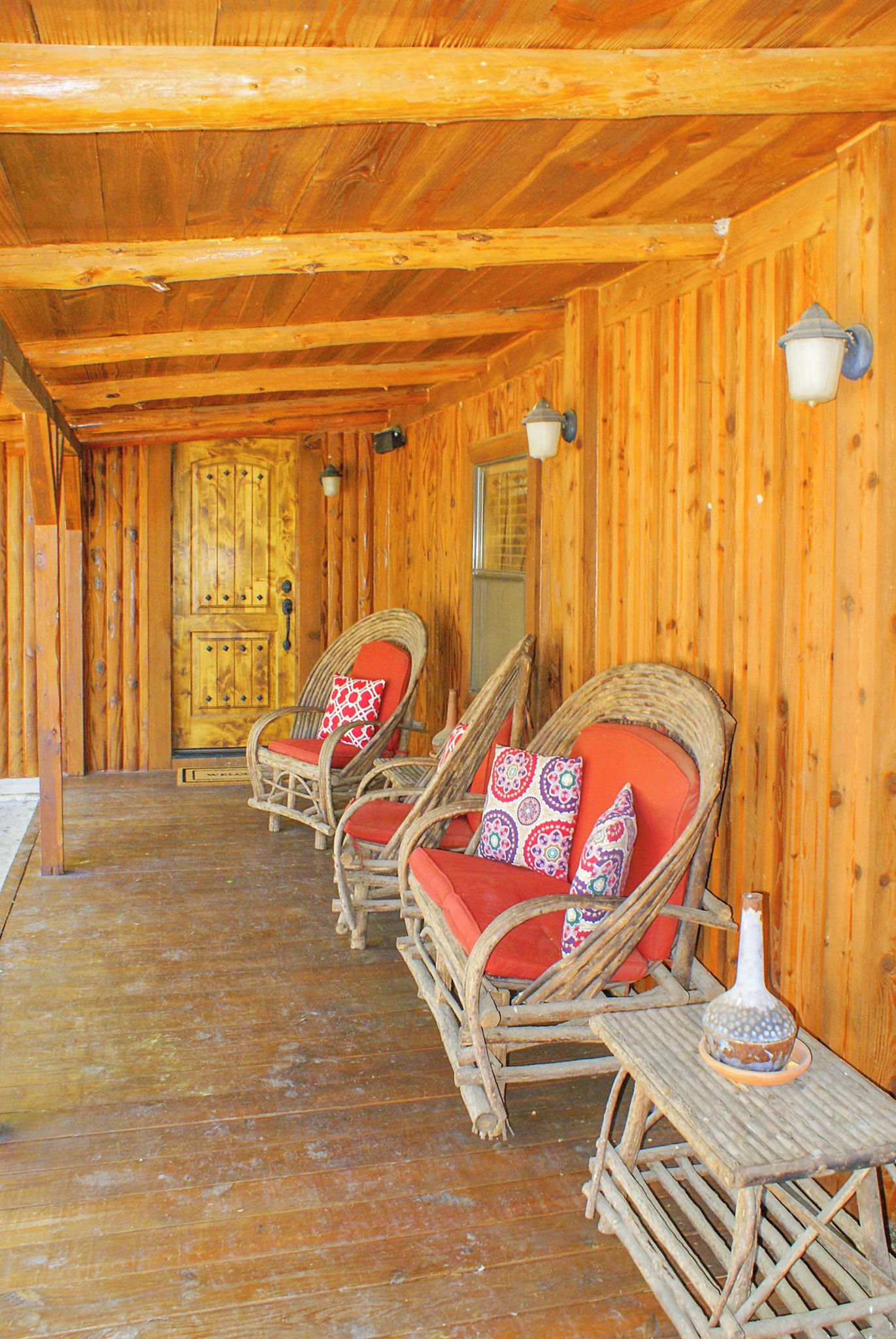 A wooden cabin with wicker chairs and red cushions, a wooden table, and a door with a window.