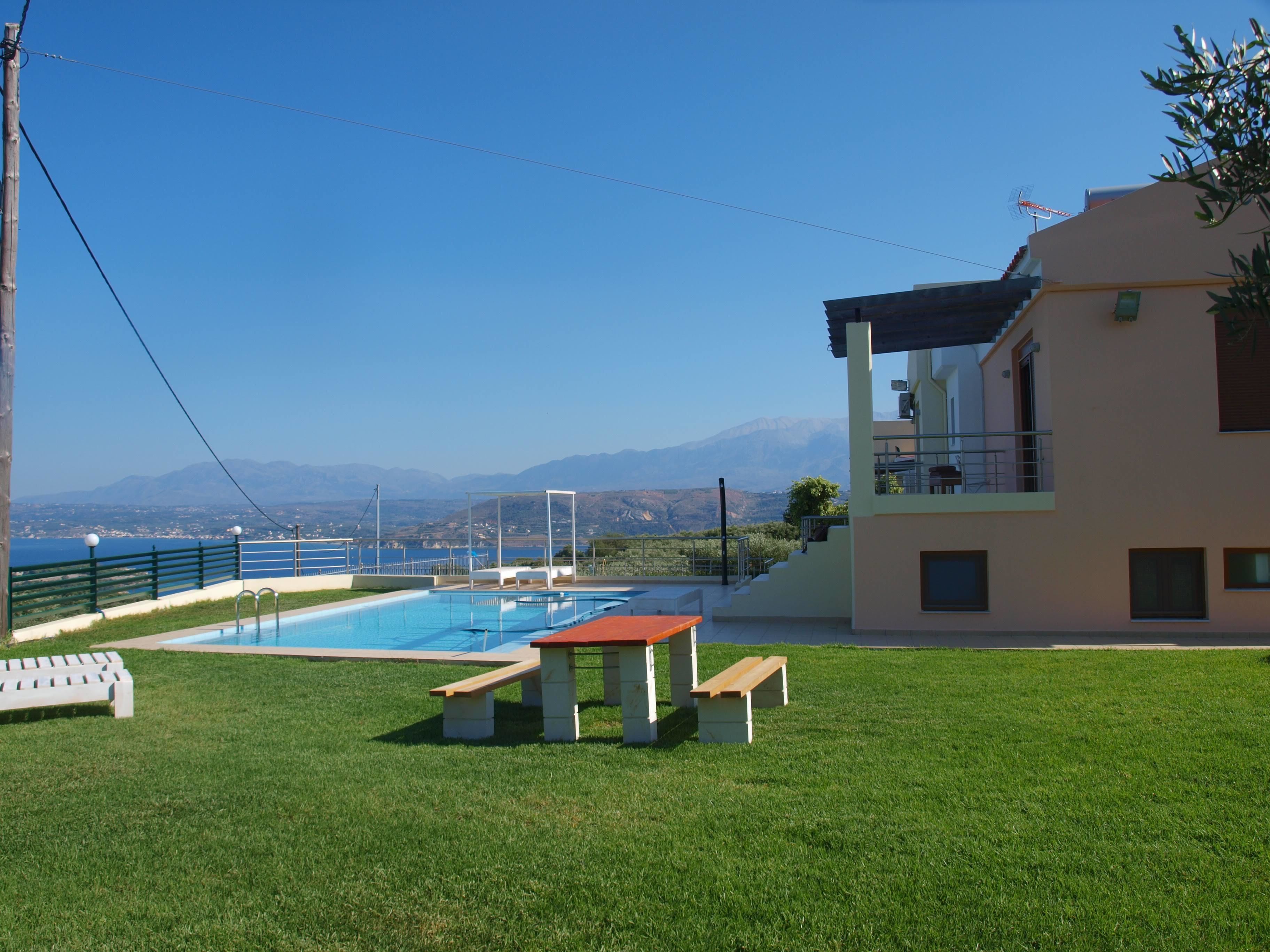 Ein Poolbereich mit Tisch und Bänken, Blick auf das Meer und ein Gebäude mit Balkon.