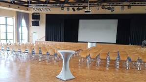 An auditorium with rows of chairs, a central table, and a stage with a screen.