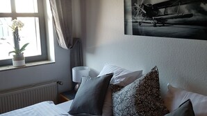 A bedroom with two beds, a window with curtains, a radiator, a lamp, and a wall-mounted airplane painting.