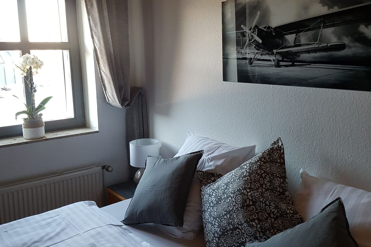 Camera da letto con due letti, una finestra con tende, un termosifone, una lampada e un quadro raffigurante un aereo appeso al muro.