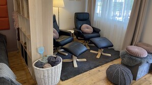 A modern living room with a black leather sofa, a black armchair, a small round table, a wicker basket with blankets, and a large window with sheer curtains.