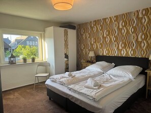 Habitación de hotel con una cama grande, una silla, un armario y una ventana con vistas a edificios y vegetación.