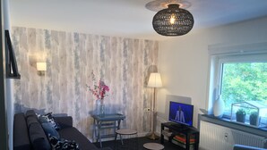 A living room with a sofa, a small table with a vase of flowers, a TV, and a window with a view of greenery. - TOP - APPARTMENT - WINTERBERG**** (max. 6 people) Prime location **** (Winterberg)