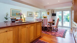 A kitchen with wooden cabinets, a granite countertop, a dining area with striped chairs, and a large window with a view of the outdoors. - Rose-Tinted Charm, by Veeve (London)