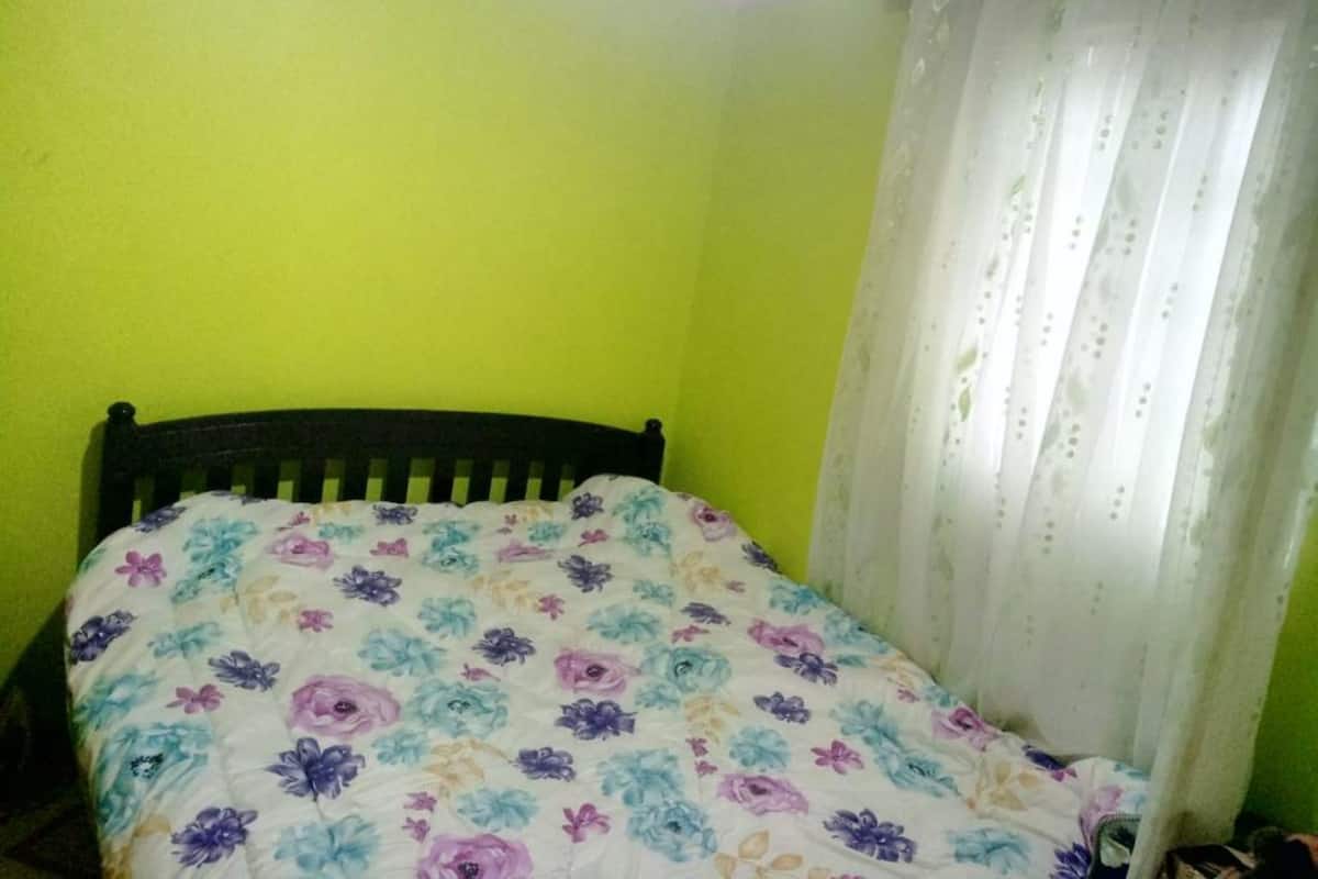 Una habitación pequeña con cama individual, colcha de motivos florales, cabecero de madera y cortina blanca.