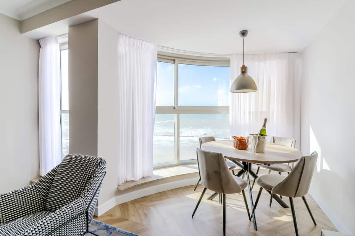 Un espace repas moderne avec une table ronde et des chaises, un luminaire suspendu et une vue sur l’océan grâce à de grandes fenêtres.