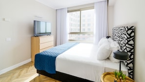 Une chambre d’hôtel moderne, équipée d’un lit, d’une télévision, d’une fenêtre avec des rideaux, d’un mur décoré d’un motif et d’une petite table sur laquelle est posée une plante.