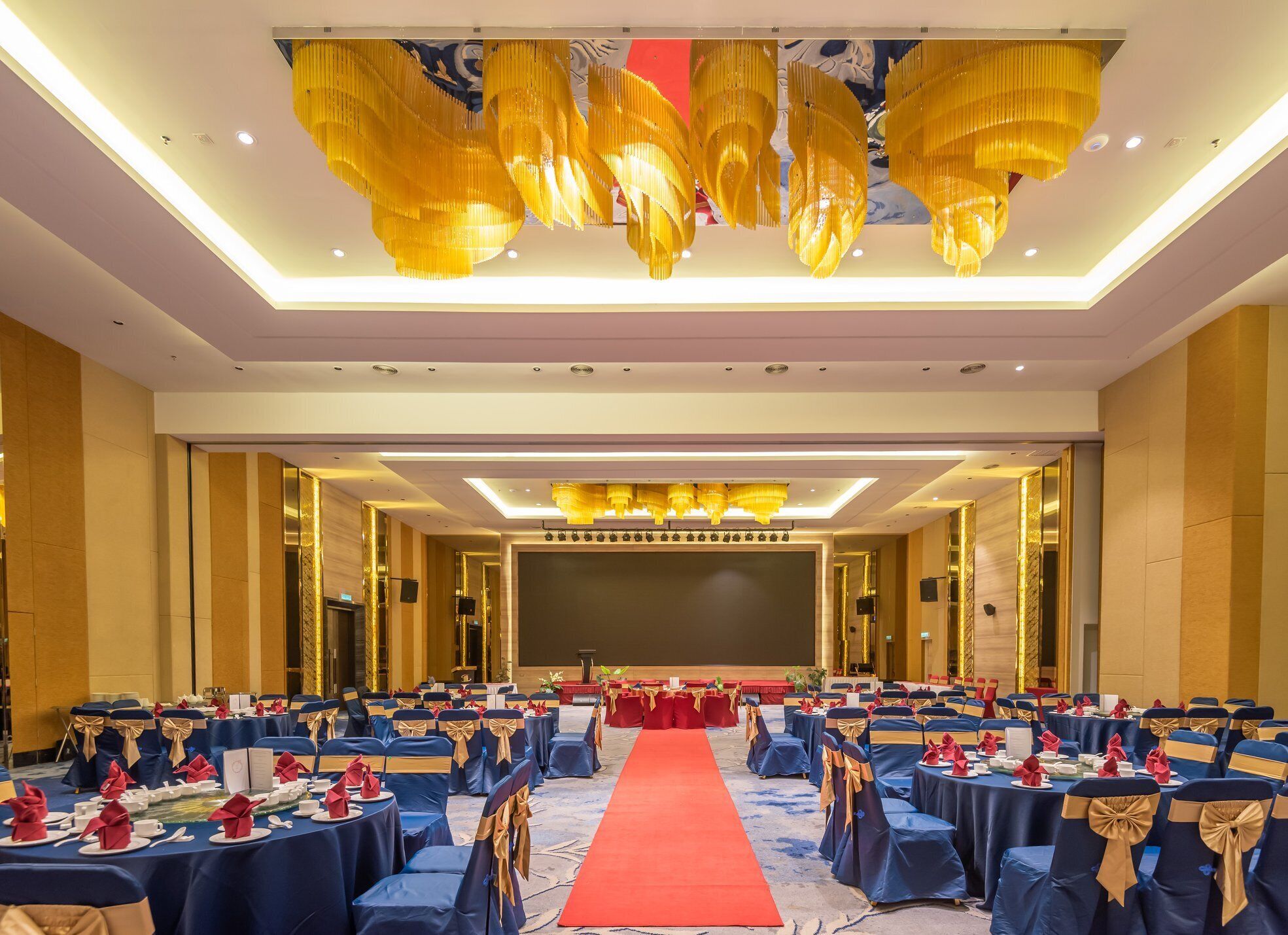 A banquet hall with rows of tables, red carpet, and golden chandeliers.
