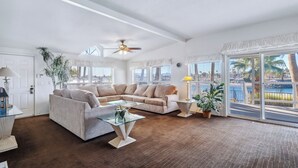 A spacious living room with a beige sectional sofa, a glass coffee table, and a ceiling fan. Large windows offer a view of palm trees and a body of water.