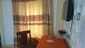 Ein Zimmer mit einem Holzbett, einem Tisch mit orangefarbener Tischdecke, einem an der Wand montierten Fernseher und einem Vorhang mit Muster.
