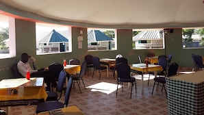 Ein Zimmer mit Tischen und Stühlen, eine Person sitzt an einem Tisch, und es gibt Fenster mit Blick auf Gebäude.