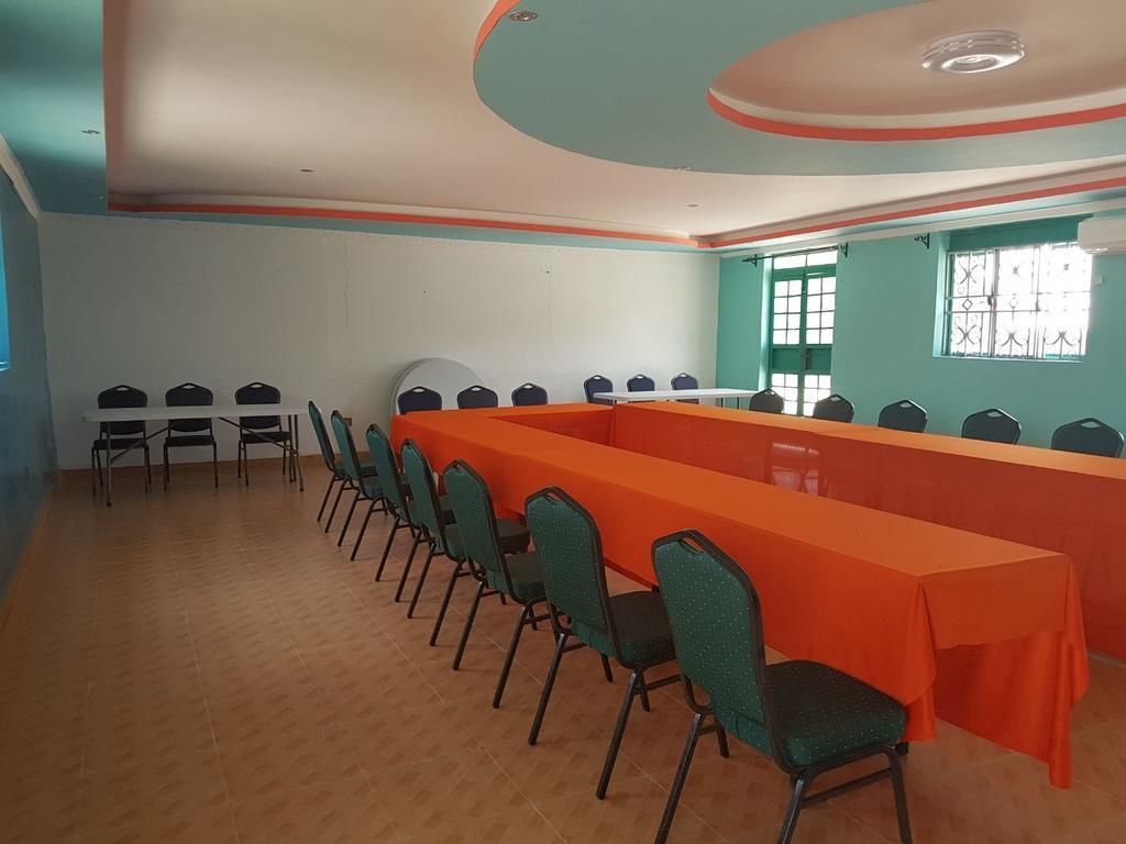 Un salón de conferencias con una mesa larga y sillas, suelo de baldosas y un ventanal con barrotes.