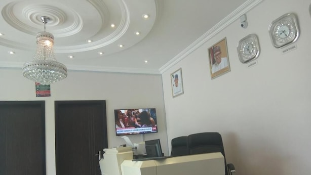 Ein Empfangsbereich mit einer Chandelüstere, einem Fernseher, der eine Nachrichtensendung zeigt, und Uhren an der Wand.