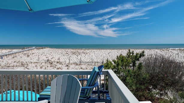Une vue sur la plage depuis un balcon, avec un parasol bleu, deux fauteuils Adirondack et une terrasse en bois.