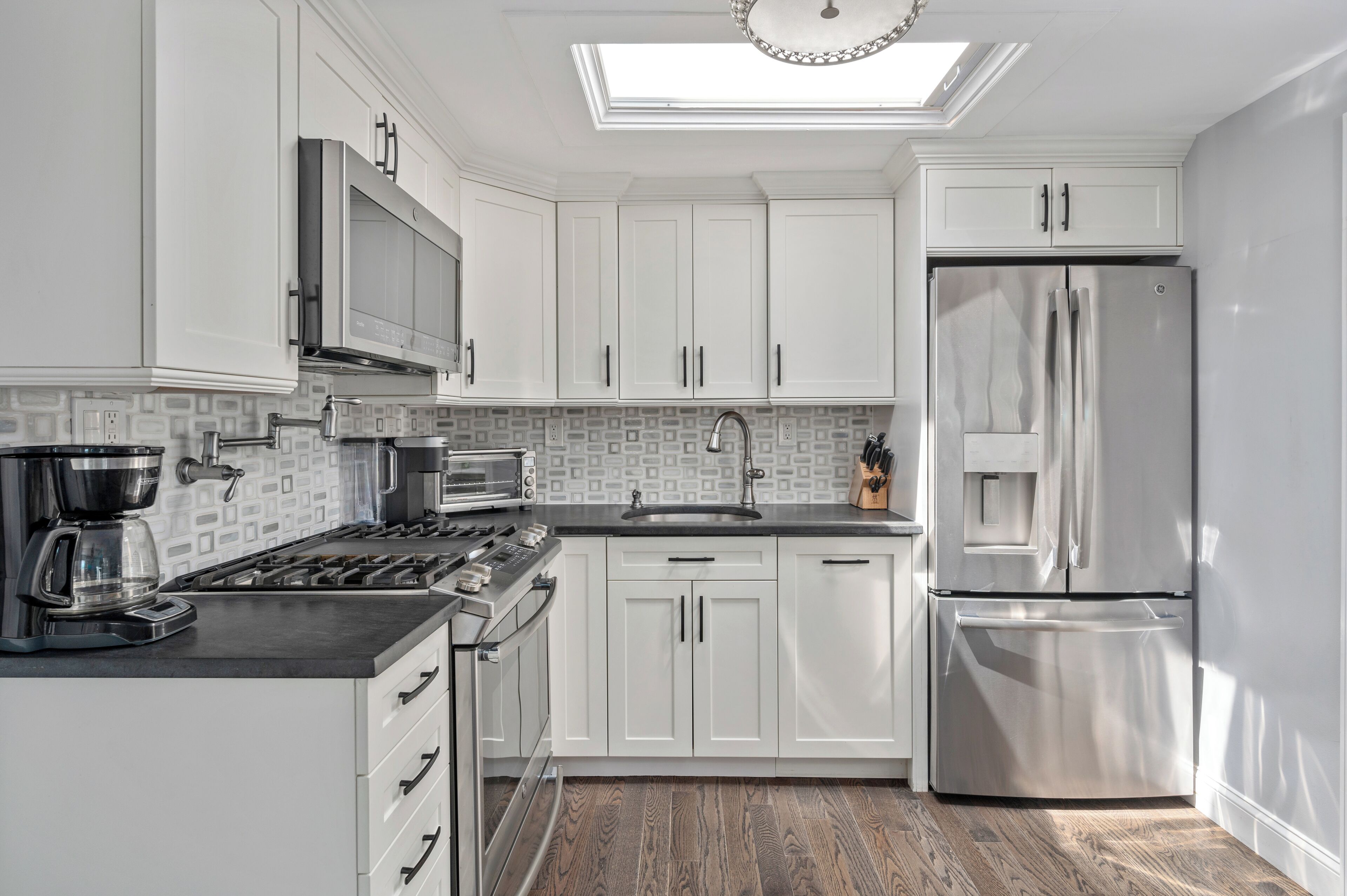 A modern kitchen with white cabinets, stainless steel appliances, a skylight, and a patterned backsplash.