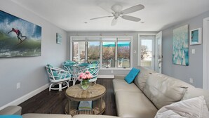 Un salon avec un canapé beige, une table basse ronde sur laquelle repose un livre, un tableau représentant un surfeur et un ventilateur de plafond.