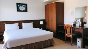 A hotel room with a large bed, a wooden wardrobe, a desk with a chair, a small stool, a lamp, and a framed picture on the wall. - Rawai Guesthouse (Chalong)
