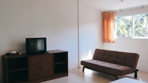 A bedroom with a bed, a TV on a wooden cabinet, a window with curtains, and a view of palm trees outside. - Rawai Guesthouse (Chalong)