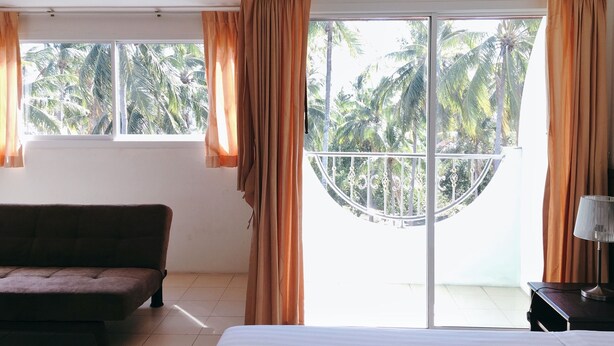 A hotel room with a bed, a sofa, a balcony with a view of palm trees, and an air conditioning unit. - Rawai Guesthouse (Chalong)