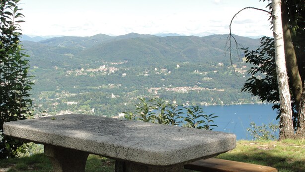 Un tavolo da picnic in pietra con panche, immerso nel verde e con vista su un lago, una cittadina e montagne sullo sfondo.