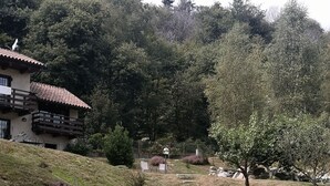 Una casa con tetto spiovente e un balcone, immersa in un prato verde con qualche albero e una recinzione.