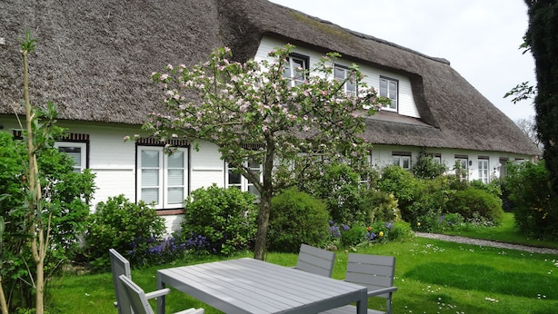 Ein Haus mit einem Reetdach und weißer Fassade, ein Garten mit Tisch und Stühlen sowie blühenden Bäumen.