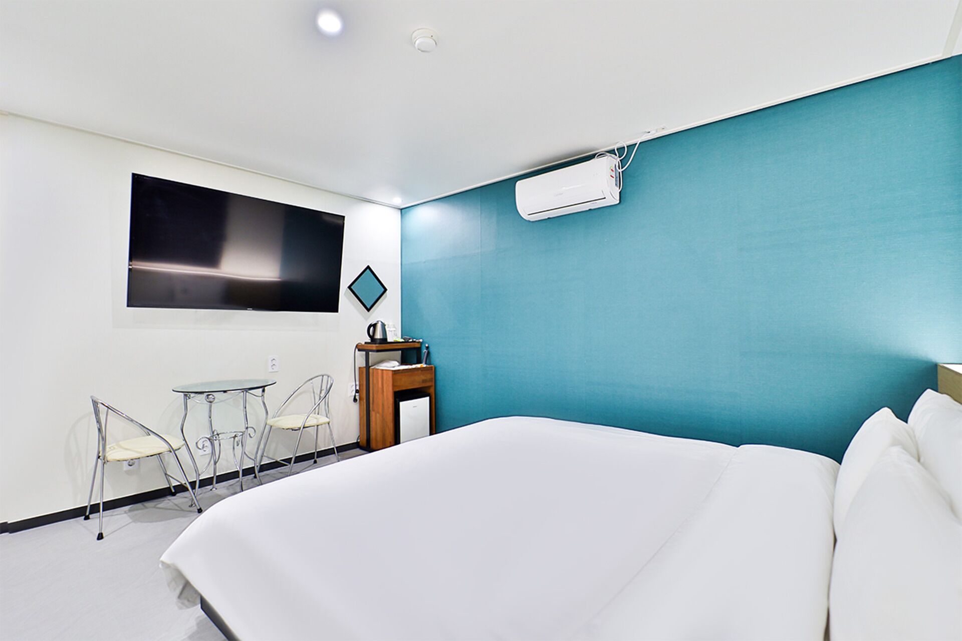 A hotel room with a bed, a desk with a chair, a flat-screen TV, and an air conditioning unit.