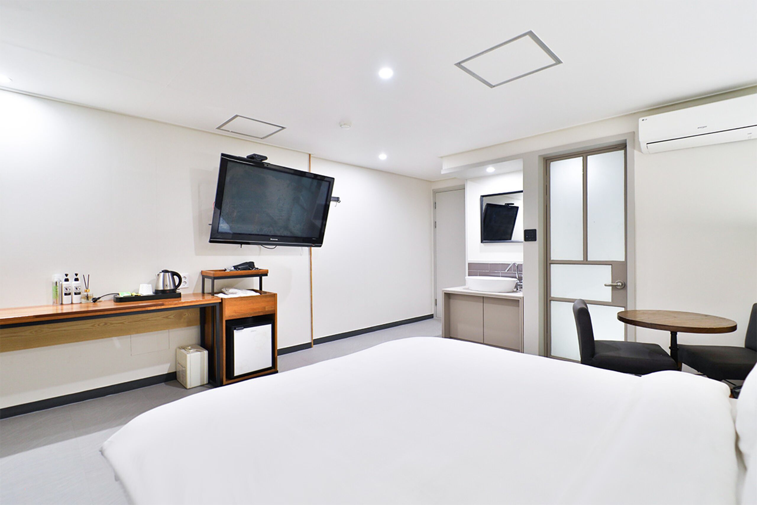 A hotel room with a bed, a desk with a flat-screen TV, a small kitchenette, and a seating area with a table and chairs.
