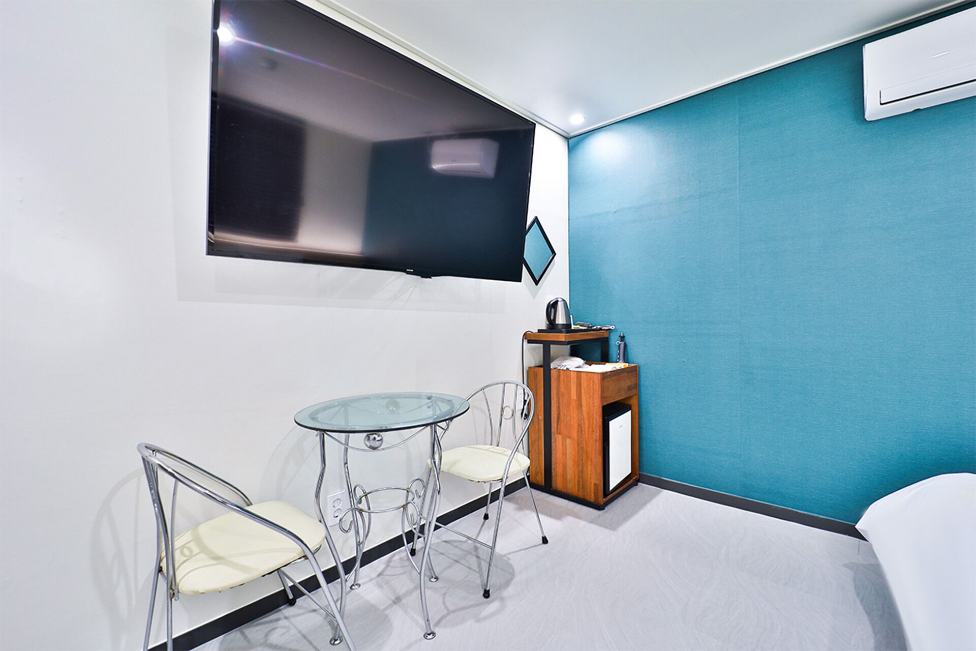 Una habitación pequeña y moderna con una cama individual, una mesita con dos sillas, un ventanal y un armario de madera con una tetera.