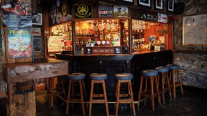 A bar area with wooden stools, a bar counter with various bottles, and framed pictures on the wall. - McGrorys (Culdaff)