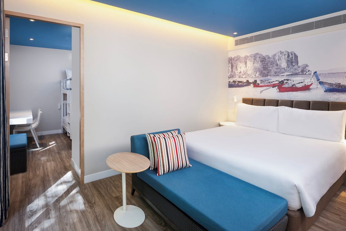 A hotel room with a bed, a blue sofa, a small round table, and a painting of boats on a wall.