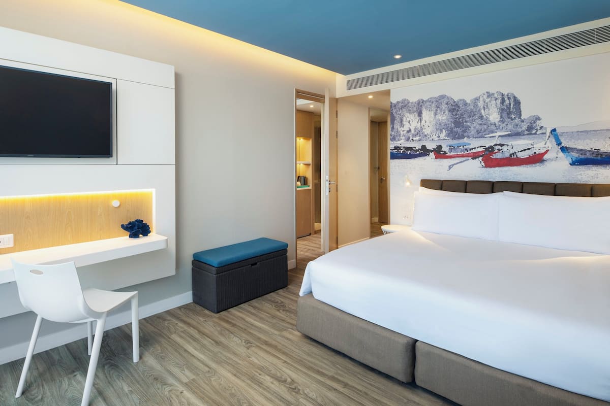 A modern hotel room with a bed, a desk with a chair, a television, and a painting of boats on a wall.