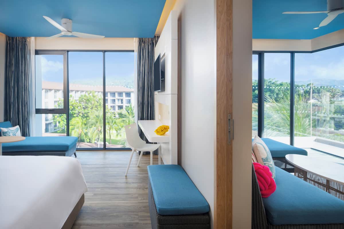 A hotel room with a large window, a bed, a sofa, a dining table, and a TV.