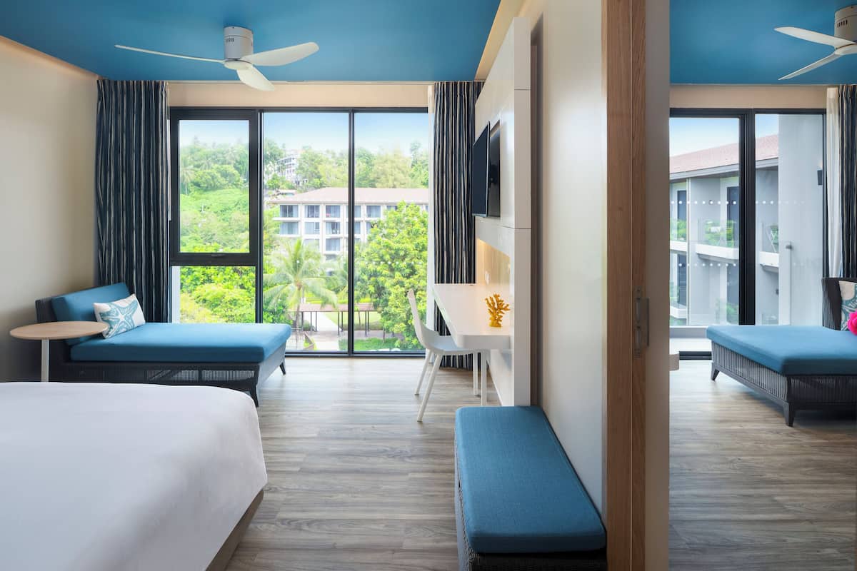 A hotel room with two beds, a desk, and a balcony with a view of buildings and greenery.